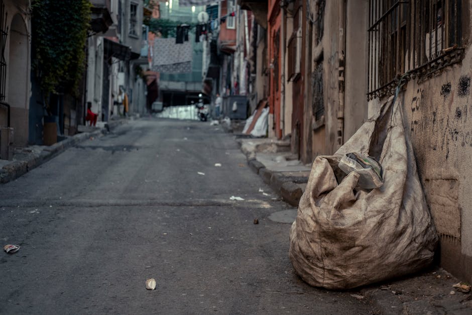 A large collection of waste materials and rubbish bags piled on a paved street corner in front of a small parking area, with a silver hatchback car parked nearby. The rubbish includes black garbage ba